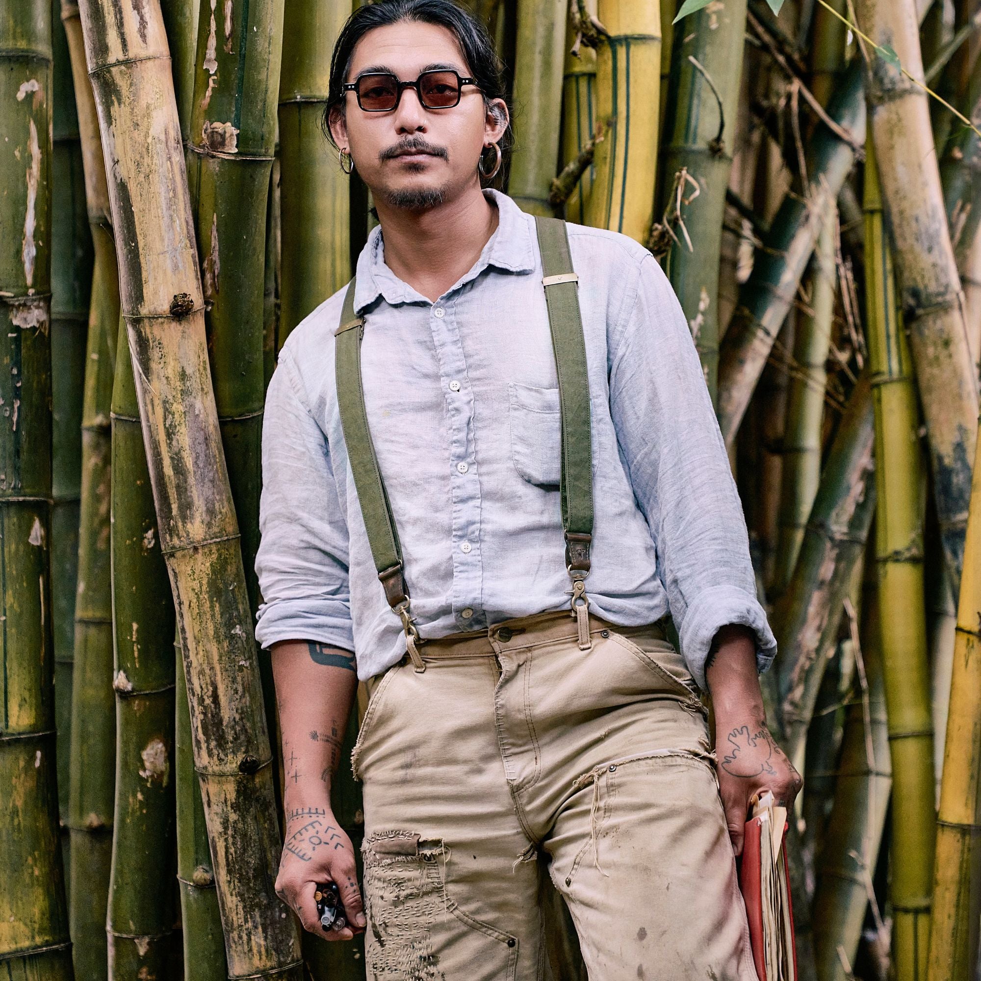 Man standing in front of bamboo wearing sunglasses, a light blue linen shirt, army green Wiseguy Original Suspenders and paints with paint marks and visible mending.