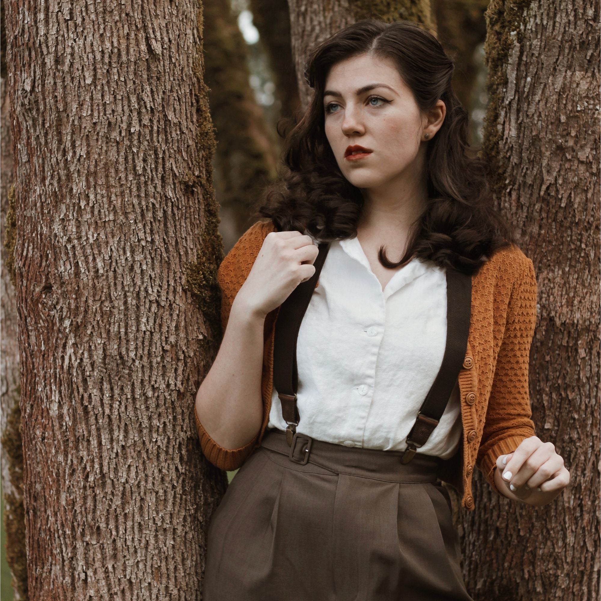 Bethany is standing in a forest, wearing a white blouse, mustard cardigan and brown Wiseguy Original Suspenders.