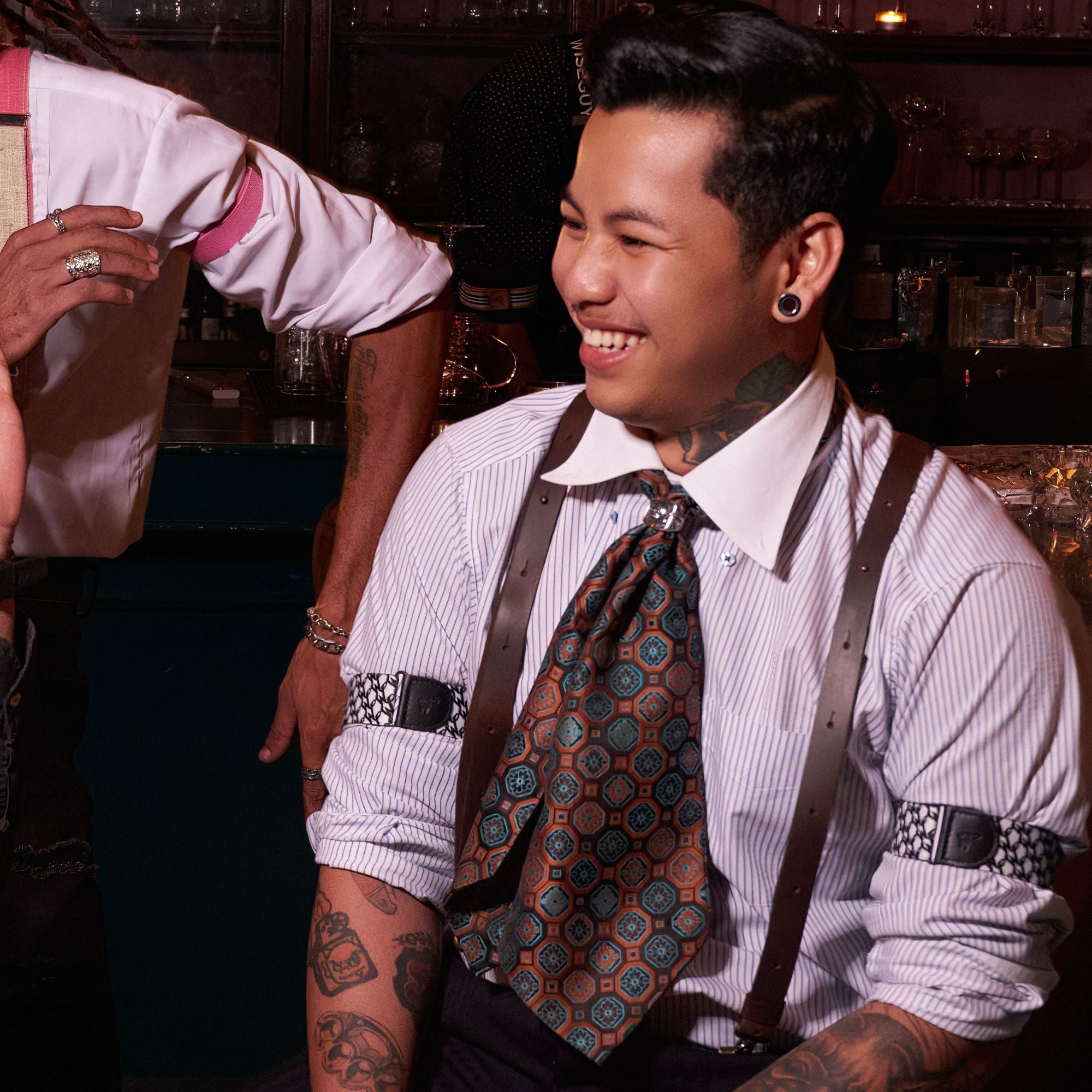 Man sitting in a bar, laughing, wearing a striped shirt with rolled up sleeves and sleeve garters. He is also wearing suspenders and a neck scarf.