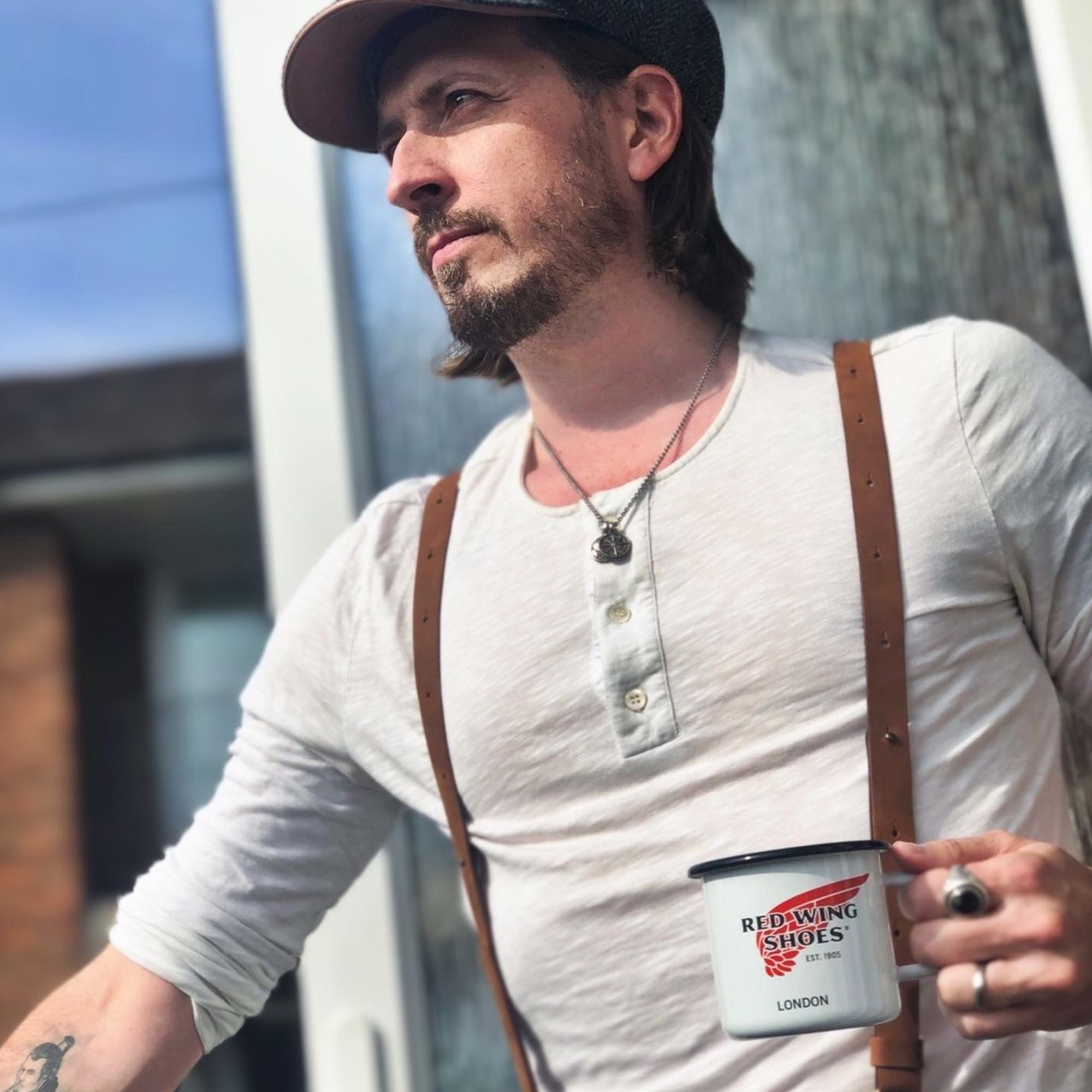 Man wearing flat cap, white henley shirt and camel brown leather Wiseguy Original Suspenders. He is holding a redwing logo coffee cup.