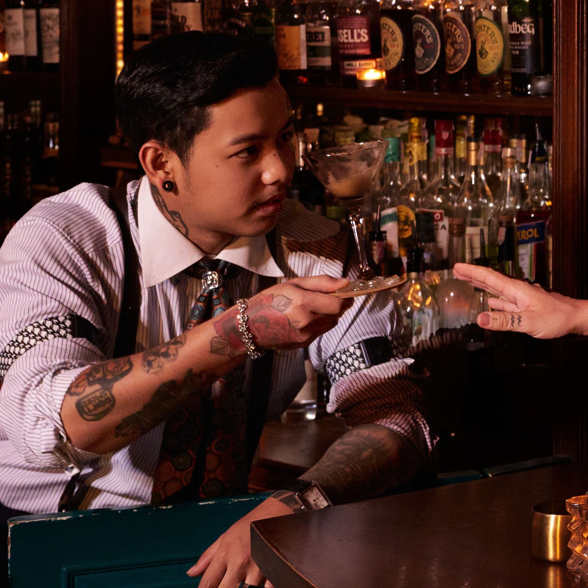 Bartender standing behind bar holding martini glass, wearing a striped shirt with rolled up sleeves and Wiseguy Original Sleeve Garters. He is also wearing suspenders and a neck scarf.