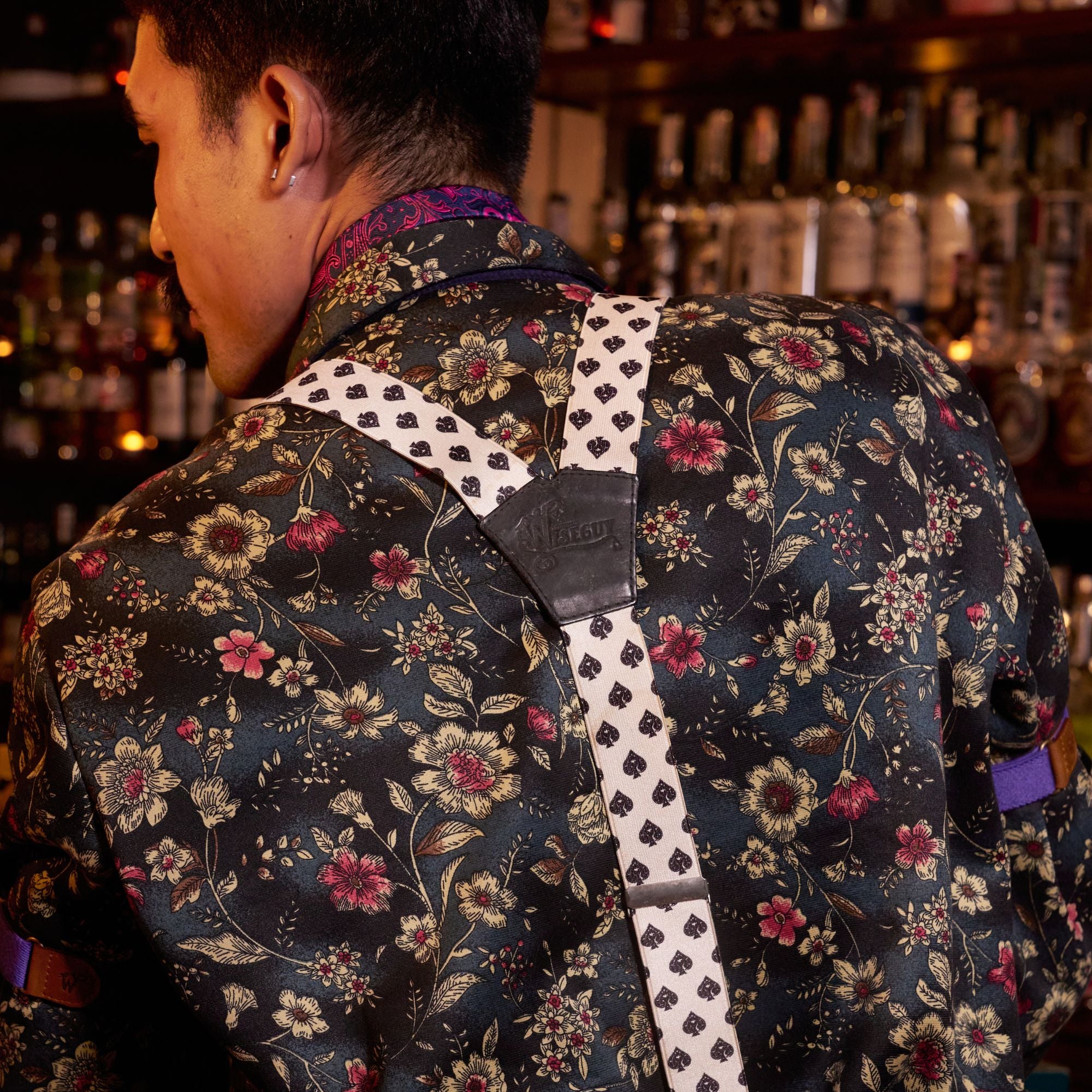 Back of person wearing patterned floral shirt, with suspenders with a spades pattern.