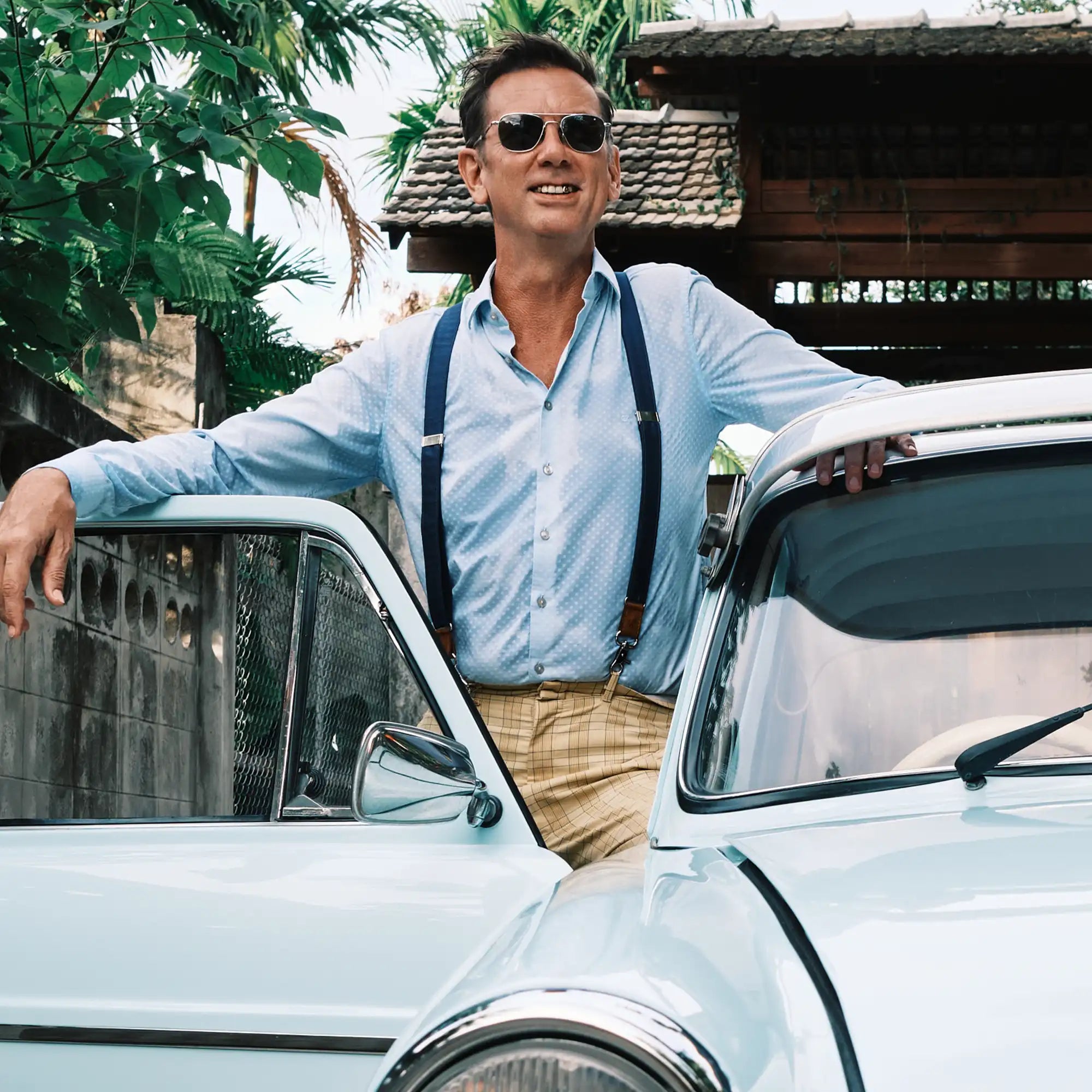 Man standing in door opening of his retro car, wearing aviator sunglasses, a light blue shirt and navy blue silk Wiseguy Original Suspenders.