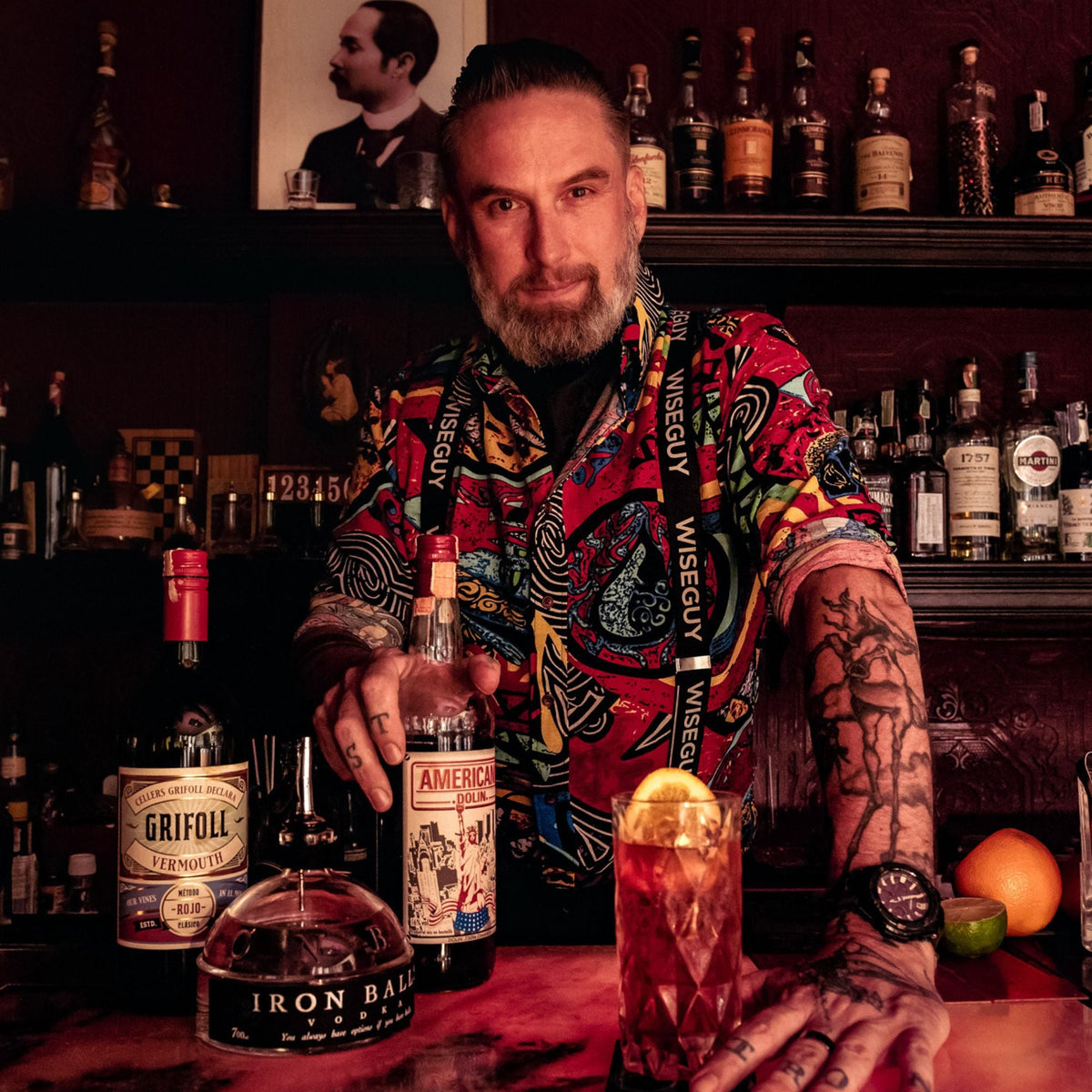 Man who is bartender with rolled up sleeves showing tattoos. He is wearing elastic suspenders with the word 'Wiseguy' on them.