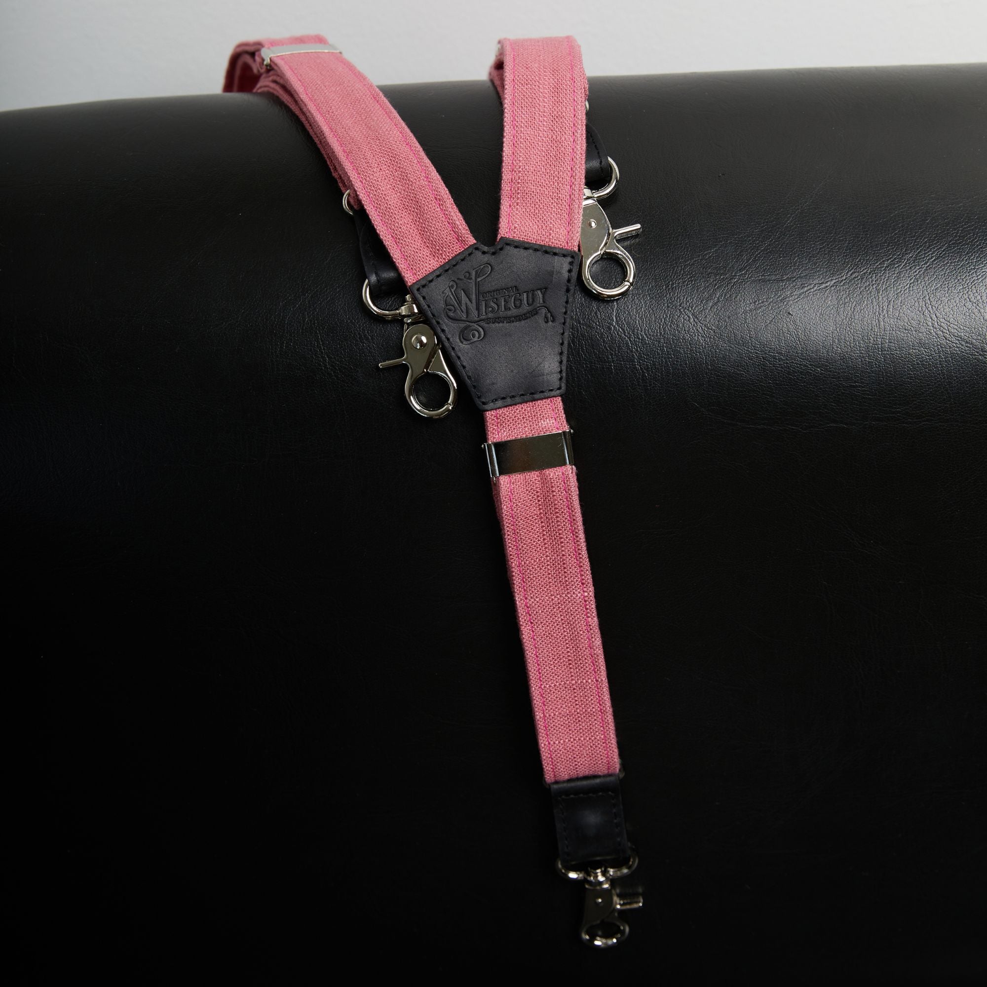 Pink linen Wiseguy Original Suspenders lying on top of a black leather couch.