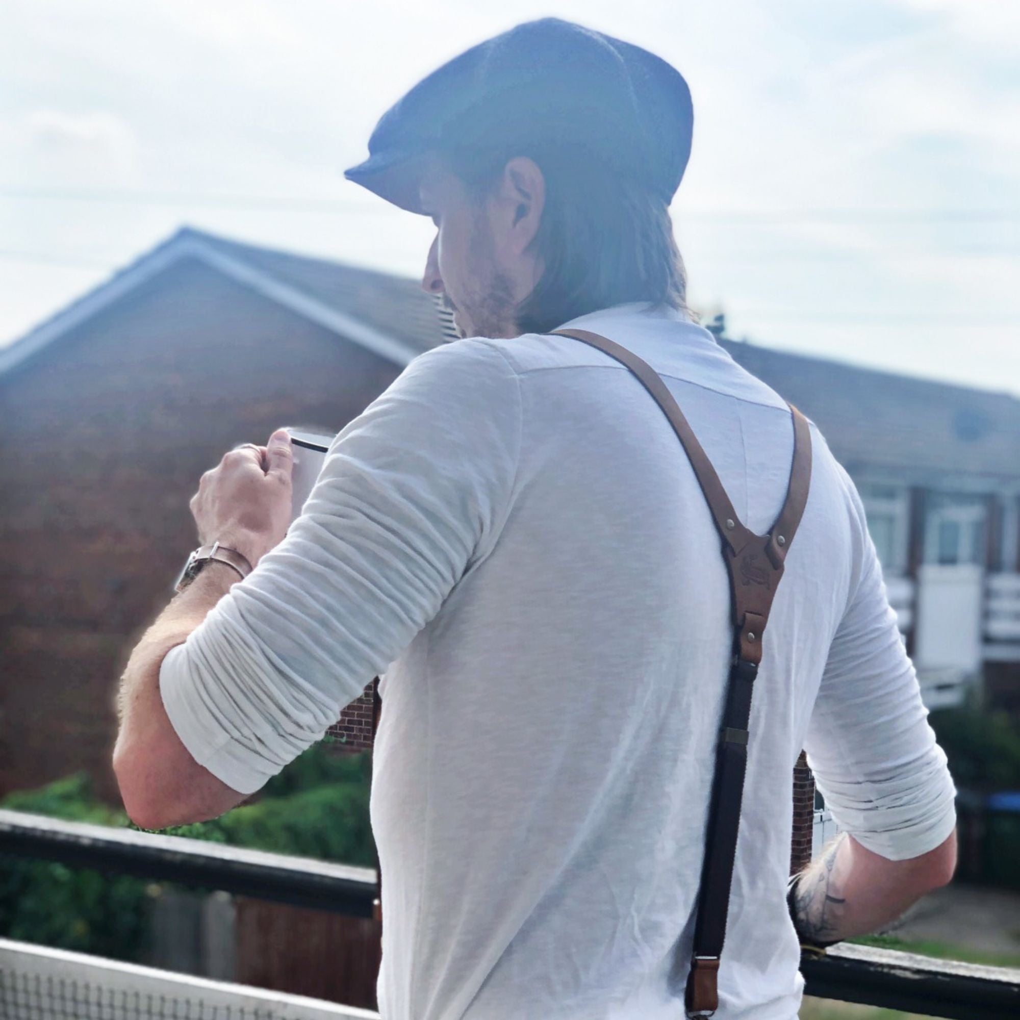 Back of man wearing flat cap, white henley shirt and camel brown leather Wiseguy Original Suspenders. He is holding a coffee cup.