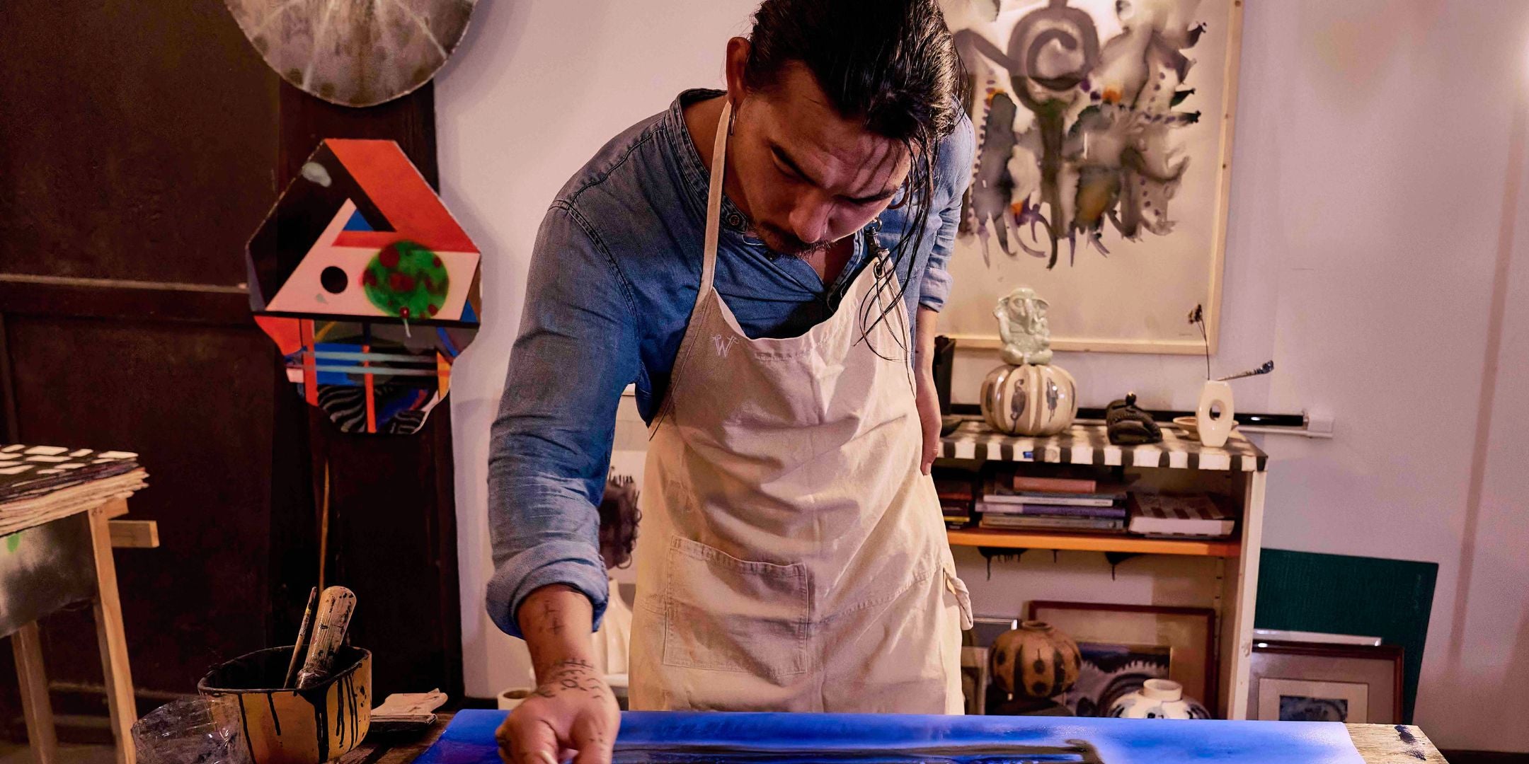 Man in an art studio painting wearing a wiseguy original creme colored canvas apron he is painting with blue and black ink