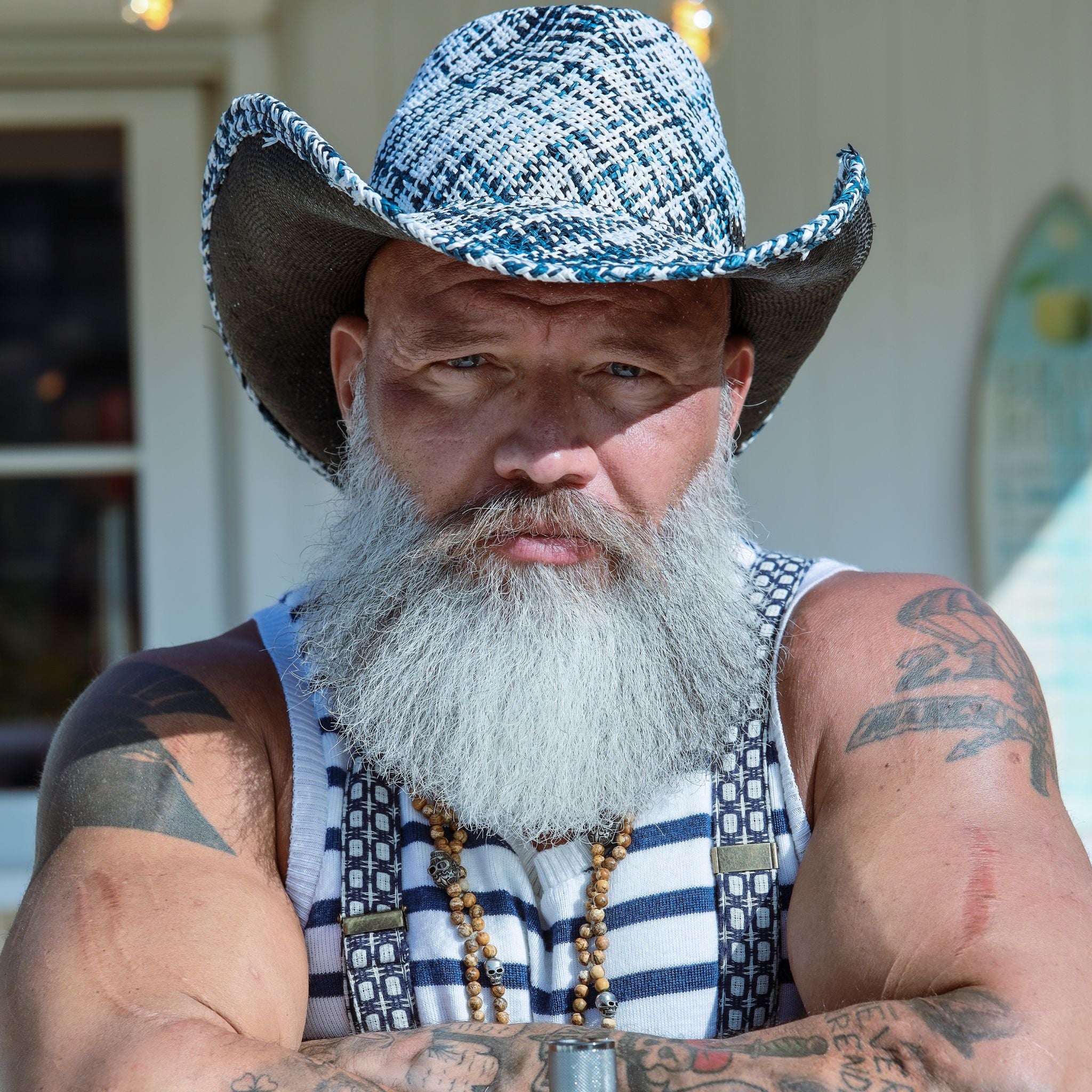 Portrait of man looking in camera, he's wearing a blue and white colored cowboy hat and checkered suspenders.